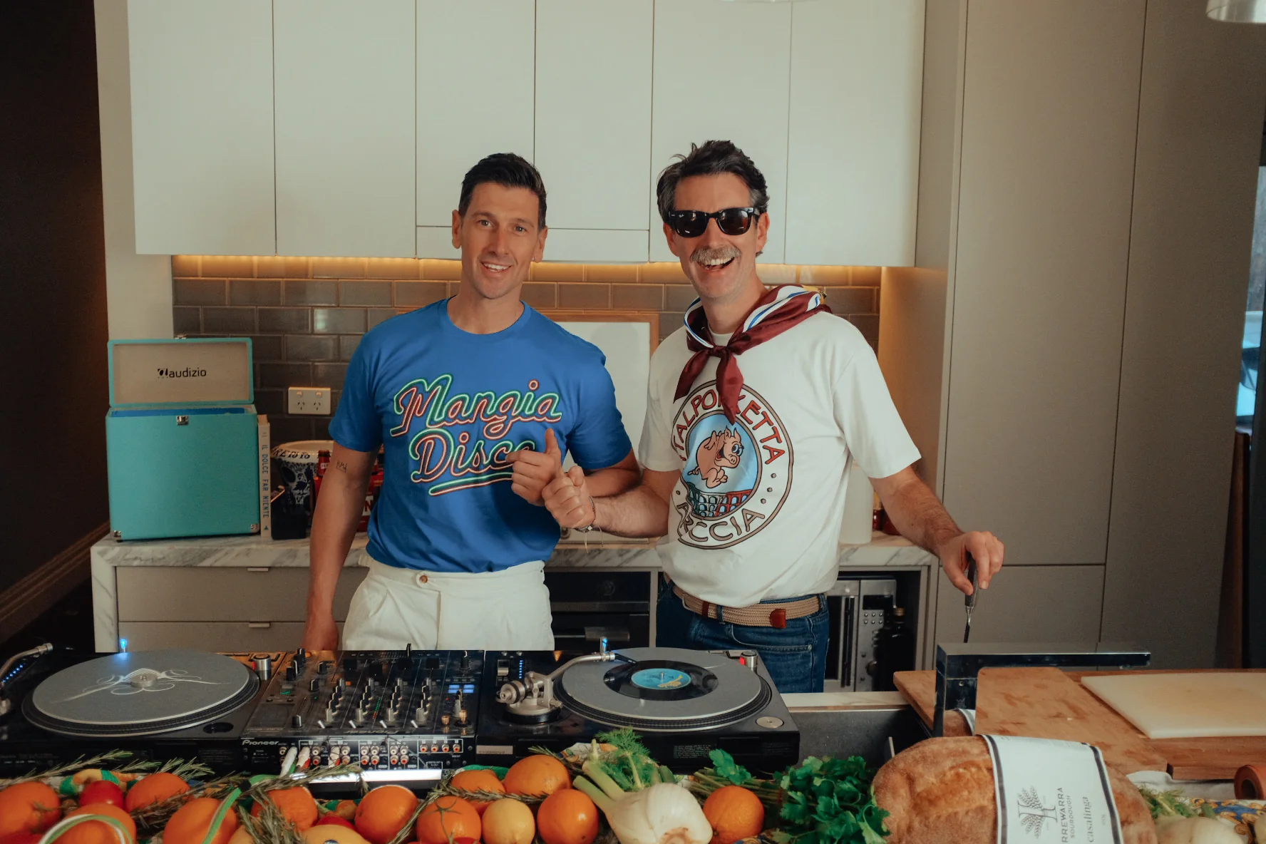 Andy in a Mangia Disco tee and Matt in a porchetta tee behind the decks, oranges and sourdough on the bench