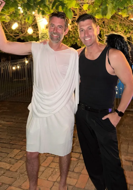 Matt in a toga and Andy in black, under festoon lights at a party